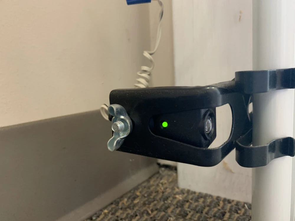 a garage door safety sensor mounted at the bottom of a vertical track near the ground. The sensor is encased in a black housing, secured with a bracket to a white support beam. A green light is visible on the sensor, indicating that it is operational and aligned correctly with its counterpart. A coiled wire connects the sensor to the garage door system. The setup appears to be part of an automatic garage door mechanism, designed to detect obstructions in the door's path and prevent accidents or damage by stopping the door's movement when triggered