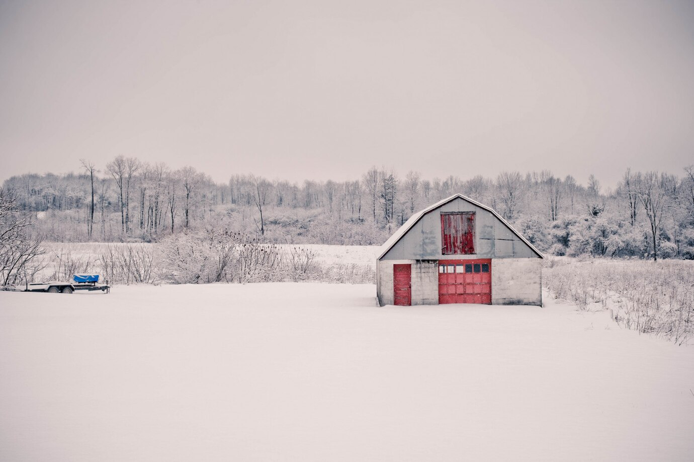 a serene, snow-covered rural scene with a barn as the central focus. The barn has a weathered gray exterior with red doors, standing out against the white blanket of snow. In the background, leafless trees form a natural border under an overcast sky, creating a calm and tranquil winter atmosphere